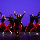 Six dancers in black tops and red pants striking energetic poses on a purple-lit stage