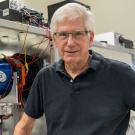 A smiling man wearing a black polo stands in front of scientific equipment.
