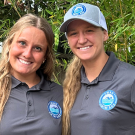 Two smiling women stand together outdoors, wearing matching gray polo shirts and caps.