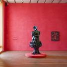 Museum gallery with red walls, wooden floor, and dark sculpture on round red plinth. Installation view: Woody De Othello: coming forth by day, Pérez Art Museum Miami, 2025–26. (Oriol Tarridas)