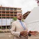 Chancellor Gary S. May in tan suit, speaks into TV camera (not seen), while stanbding in front of Aggie Square building