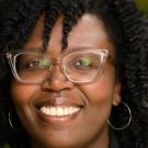 A woman wearing a black blazer smiles in front of a background of greenery