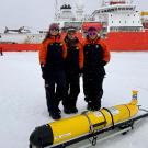 Three scientists in orange jackets stand on snow with a yellow underwater drone and a research ship behind them.