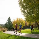 Two joggers on a sunny park path with autumn trees and two walkers nearby