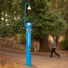 Person walks past blue emergency call station among redwood trees.