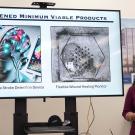 Two people stand and speak in front of a screen that reads "screened minimum viable products," showing renderings of medical devices