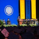 Graduation stage with blue-and-gold backdrop, university seal, speakers and mortarboards