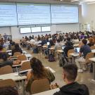 Seen from behind, students in tiered seats face three large screens at the front of a large lecture hall.