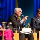 Three speakers engaged in dialogue on stage, with a blue backdrop and plants visible.