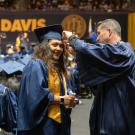Smiling UC Davis graduate in cap and gown having tassel adjusted by staff volunteer