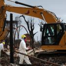 Two workers in white hazmat suits beside a yellow excavator at a muddy, bare-treed site