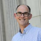 Smiling man with glasses in blue shirt standing by stone columns