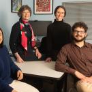 Four people smiling in an office setting, standing and sitting around a desk.