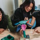 Three young women collaboratively examining a colorful, plant-like robotics project on a table.