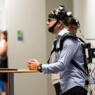 Person wearing head-mounted tracking gear and backpack, leaning on a table in a hallway.