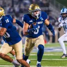 Football players in blue and gold; ball carrier runs as teammate blocks, white defender pursues