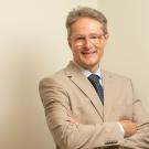 Smiling man in beige suit and blue tie, arms crossed against neutral background