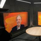 Chancellor Gary S. May, in suit and tie, at round table with microphone, in studio, for "Face to Face" recording with Bob Segar, on a monitor, appearing remotely from Boston