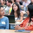 Three students conversing in a crowded lecture hall, laptops and notebooks on desks
