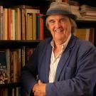 Smiling man in hat and blue jacket by bookshelf, warm-lit photograph.