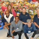 Seven employees pose in front of tables stacked with food donations.