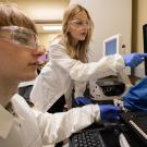 Two research students in lab coats and safety goggles examine a green display on a computer screen.