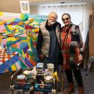 Two people pose in an art studio with a colorful abstract painting in the background.