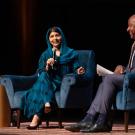 Malala Yousafzai, seated on stage, holds a microphone and speaks while Chancellor Gary S. May, seated nearby, laughs