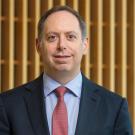 A man in a navy suit and red tie stands in front of a wood-paneled background 