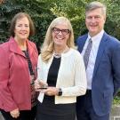 Three people pose for photo outdoors. One is holding an award.