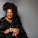 Smiling woman with curly hair wearing a black feathered coat and sparkling bracelets, gray backdrop
