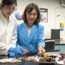 In a laboratory setting, an agricultural engineering student in a white lab coat works with their Professor, to their left, in a blue coat. They both are smiling and enjoying working together.