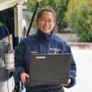 A woman holds a computer outside a bus's rear panel that is open