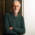 A man with glasses and a beard smiles while standing in a hallway with office doors.