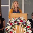 Woman speaking at podium adorned with red, white, purple floral wreath; two seated attendants