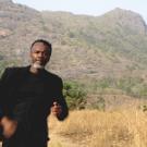 Photo of a man with brown skin in a dark suit looking at camera. He is standing in a field of tall golden grass and there are low mountains in the background.