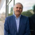 Man with gray hair and mustache in blue suit leaning against glass wall