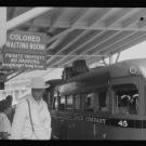 A historical black-and-white photo of a waiting area with a bus and a sign indicating "Colored Waiting Room."