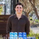 Smiling man outdoors behind table of blue-and-white milk cartons and coffee maker