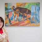 A woman in a red shirt and beige vest stands beside a colorful dog painting in an art gallery.