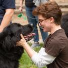Person crouching on grass, smiling while petting large black dog in red harness