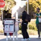 One person drops their ballot into a metal drop box while another takes a photo. A third person waits to drop off their ballot.