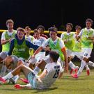 Youth soccer players cheering on lit field at night; goalkeeper on ground