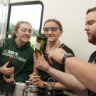 A Halsey Bottling employee holds up a wine bottle and points to it while explaining a point to two smiling students in safety goggles.