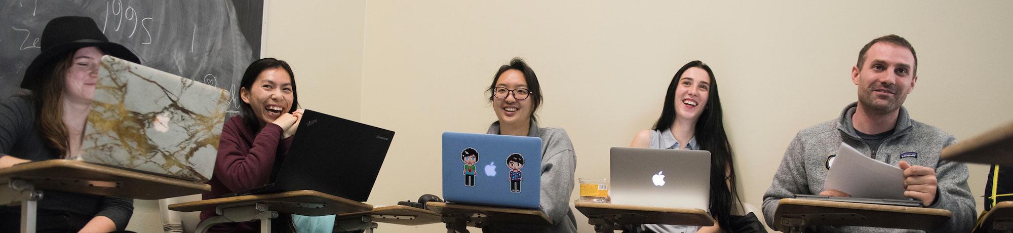 Students sit at desks smiling behind their laptops