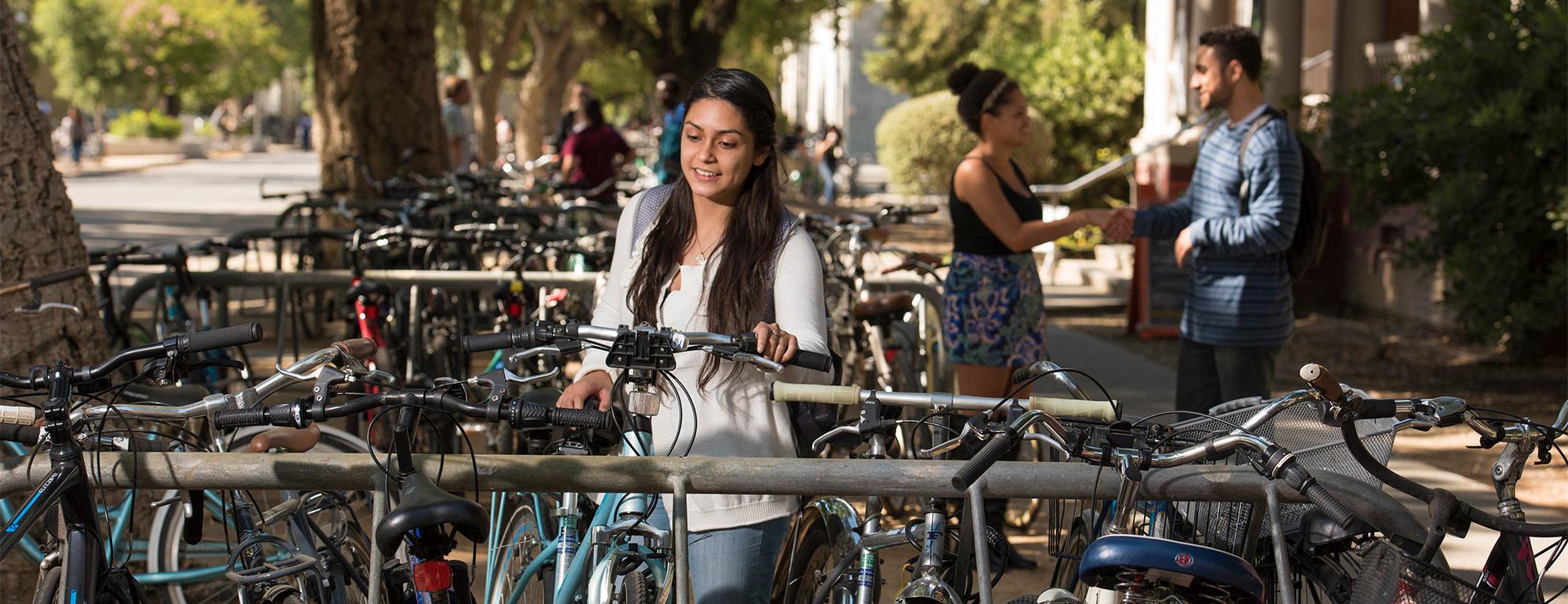 a student unlocking bike in a bike rack full of bikes and two students in the background talking