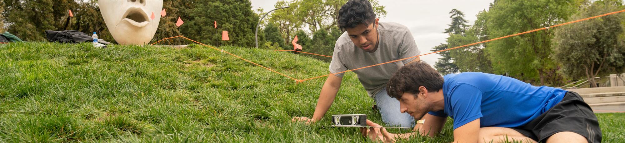 Two male students survey a landscape site with one of the UC Davis eggheads in the background