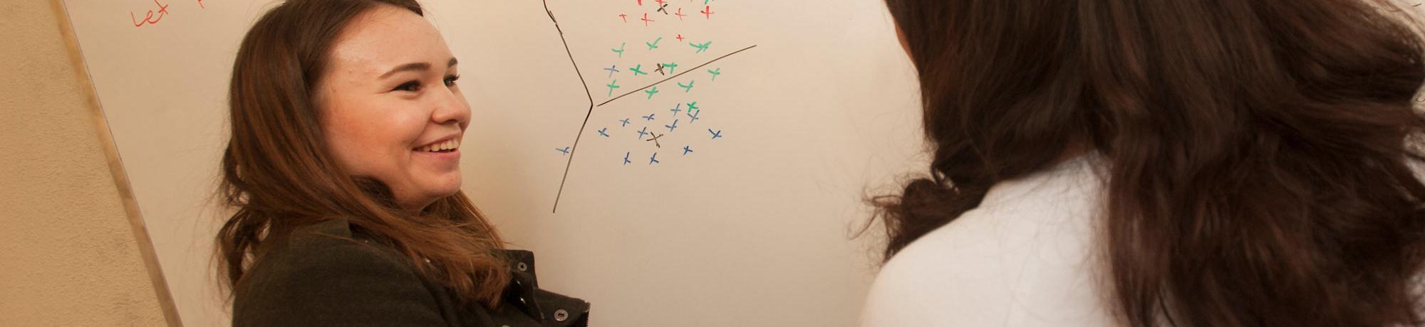 Two female students discuss a math problem at a whiteboard