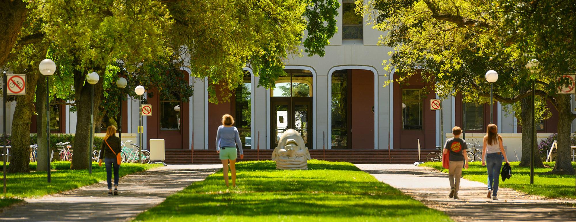 Four people walking in front of Mrak Hall on campus. One of the Egghead statues 'Eye on Mrak' is in the background.