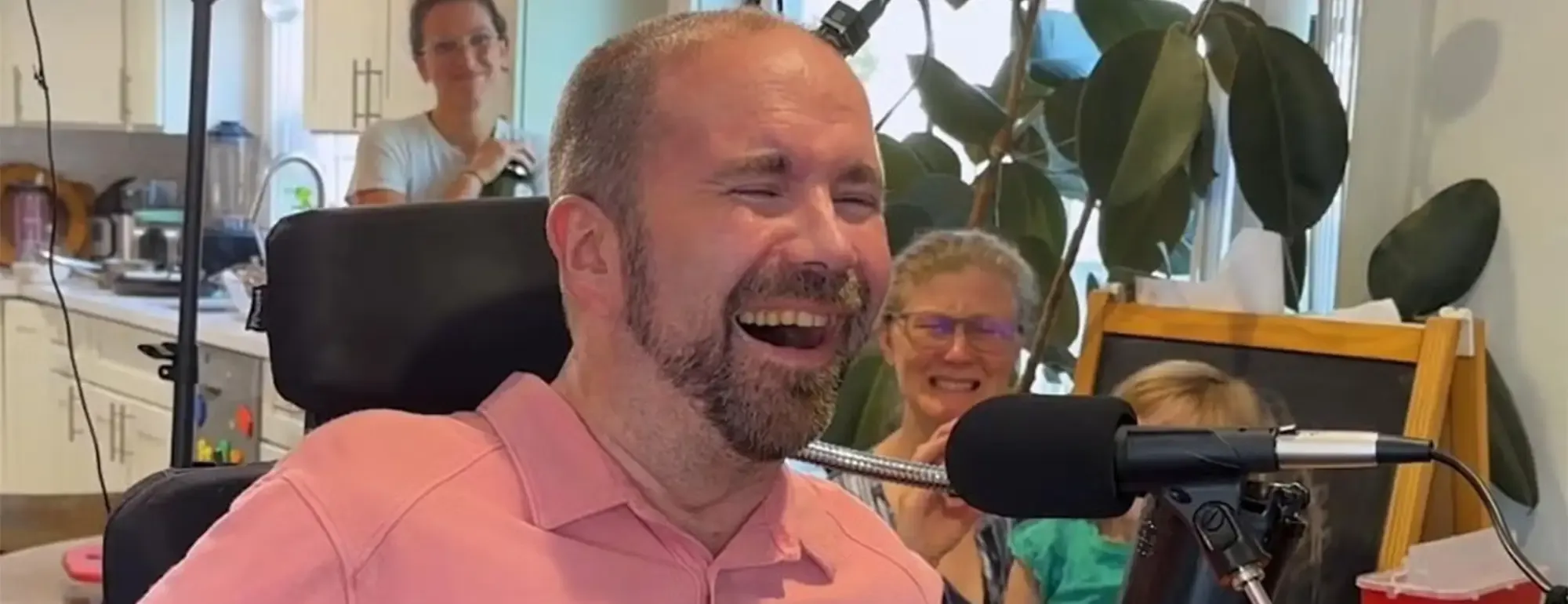 Man in wheelchair wearing pink shirt, laughing into microphone in bright office; person behind.